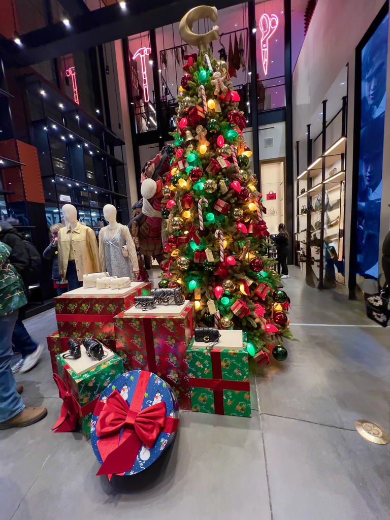 Festive store display featuring a large Christmas tree adorned with colorful lights and ornaments, surrounded by mannequins and wrapped gift boxes.