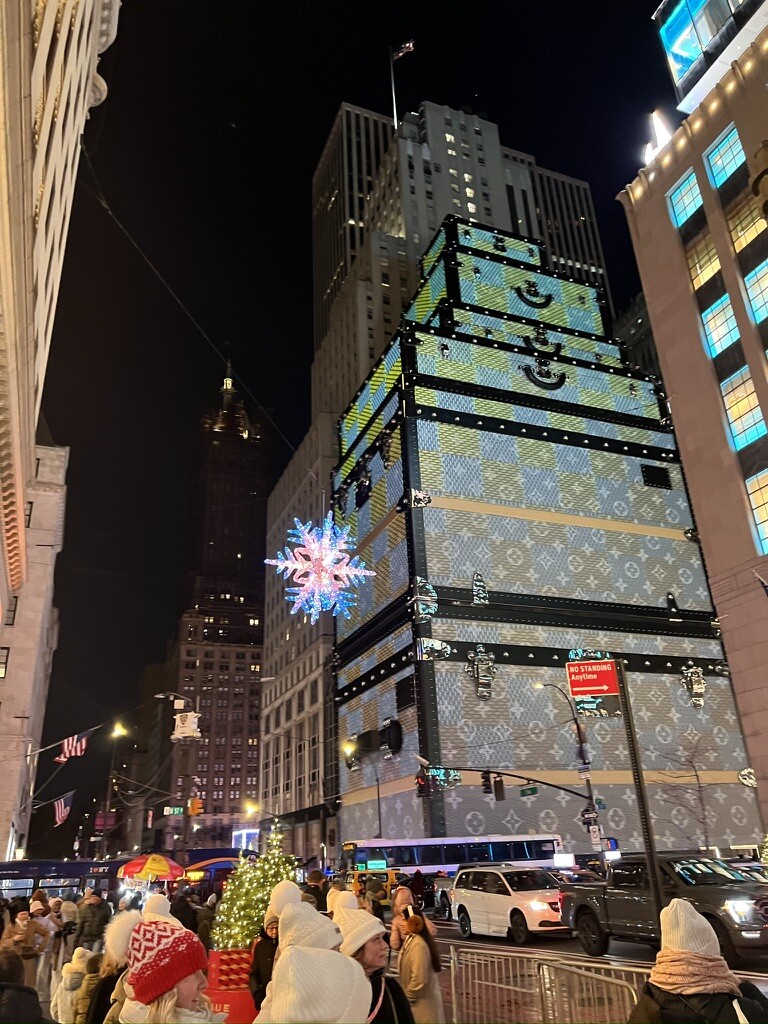 A festive city street at night, featuring a building wrapped like a giant gift with a snowflake decoration. Crowds in winter attire add a cheerful vibe.