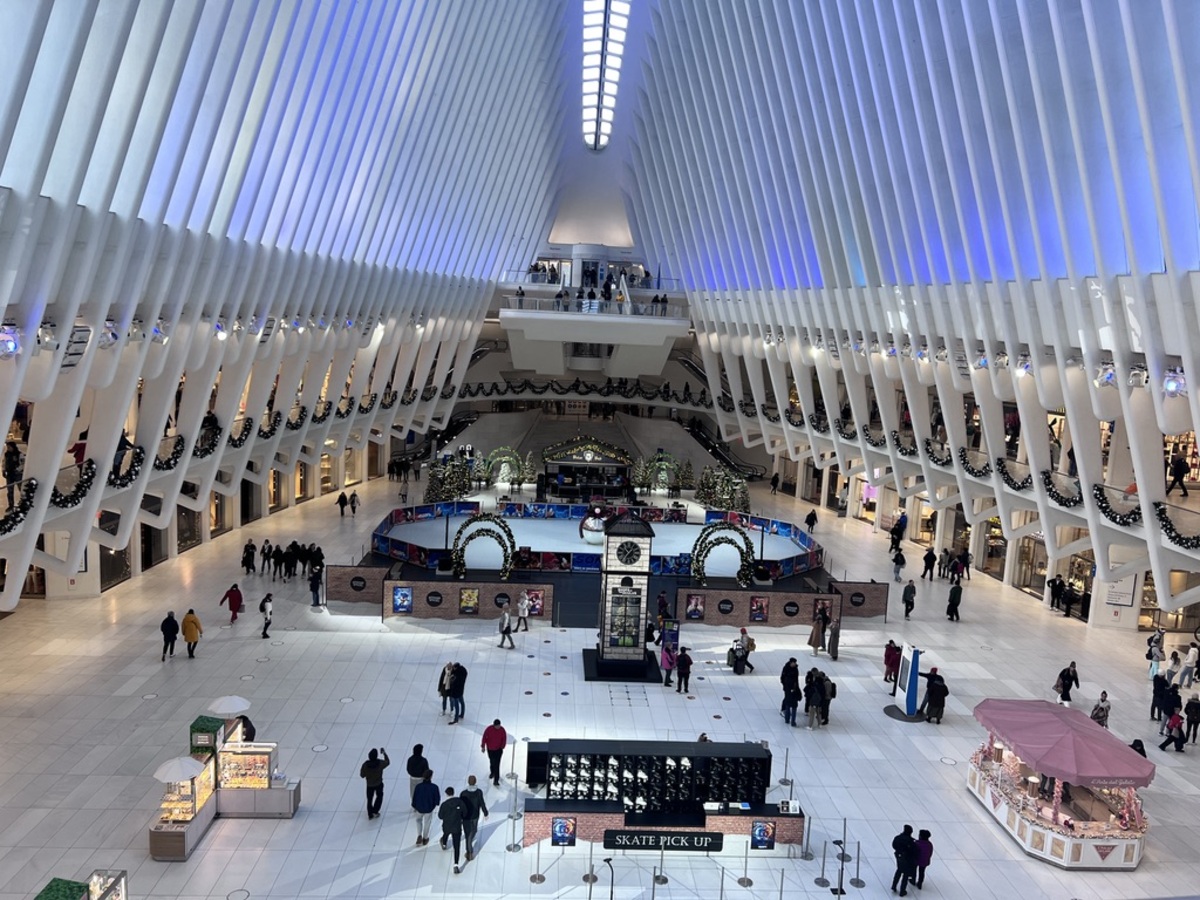 Spacious mall interior with a vaulted ceiling, blue and white lighting, and holiday decorations. People stroll by a central ice rink, creating a festive atmosphere.