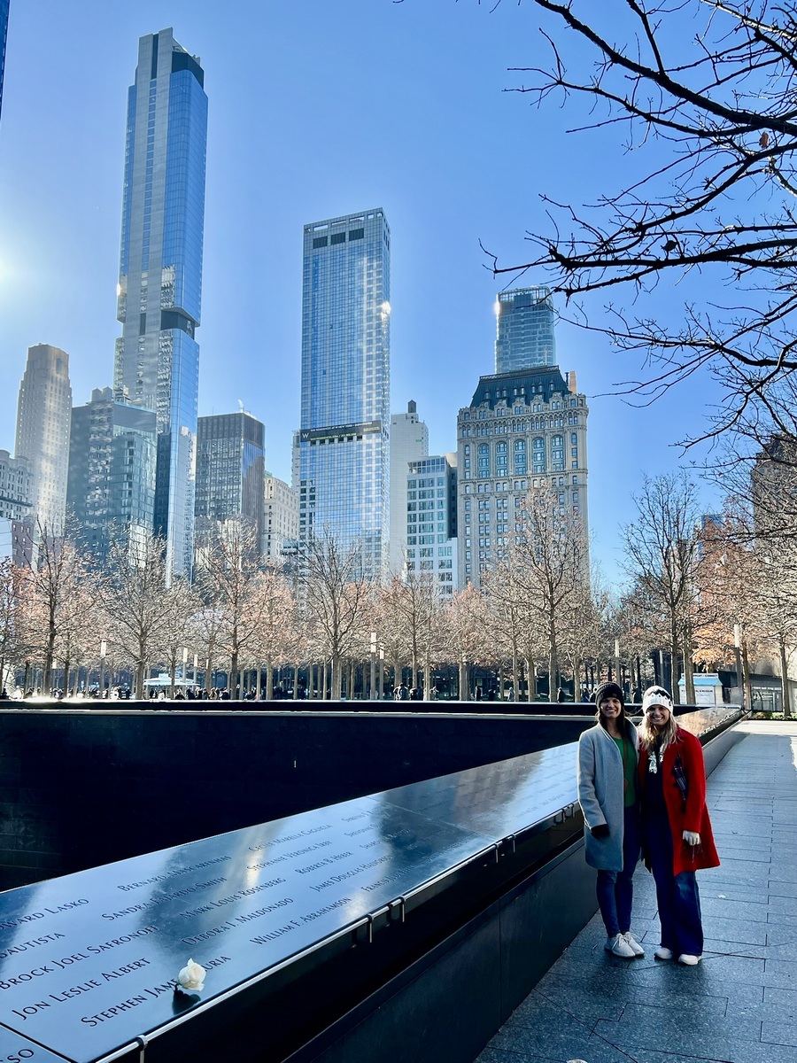 Two people stand smiling near the 9/11 Memorial in New York, with reflective panels bearing names. Skyscrapers rise in the clear blue sky, creating an urban backdrop.