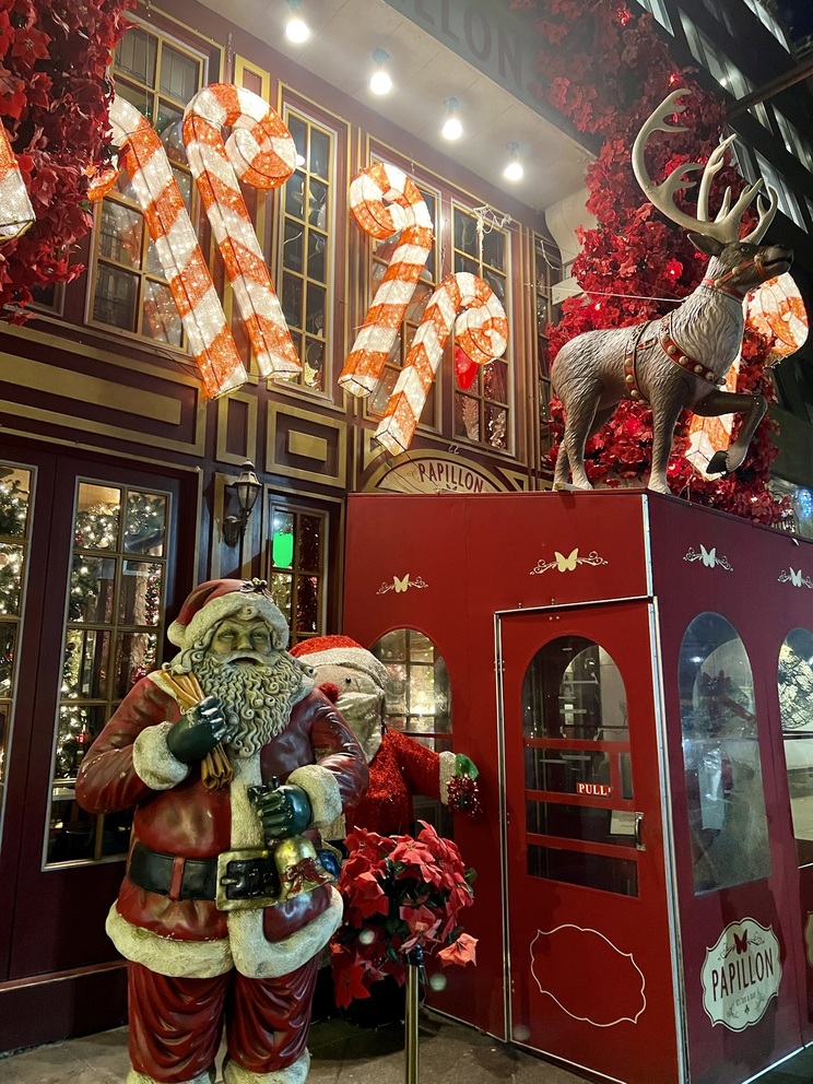 Festive storefront with a Santa statue, illuminated candy canes, and reindeer. Red and gold tones, conveying a joyful holiday atmosphere.