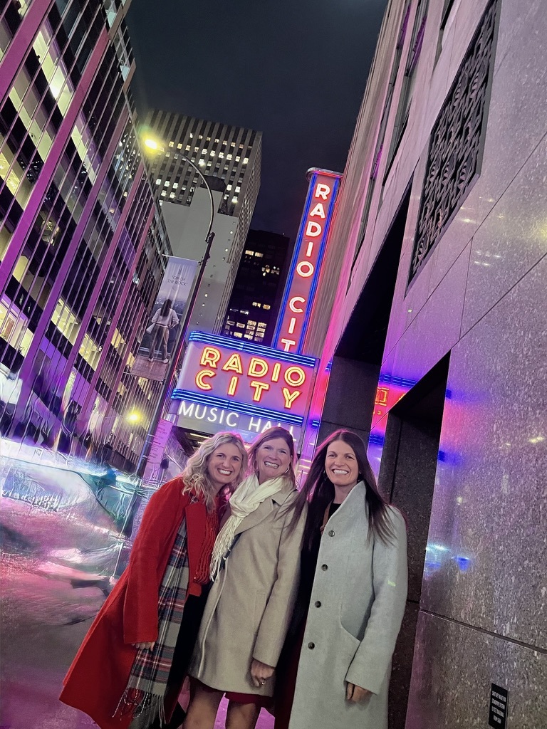 Three women smile in front of the illuminated Radio City Music Hall sign at night. Bright neon lights reflect on the wet sidewalk, creating a vibrant city atmosphere.