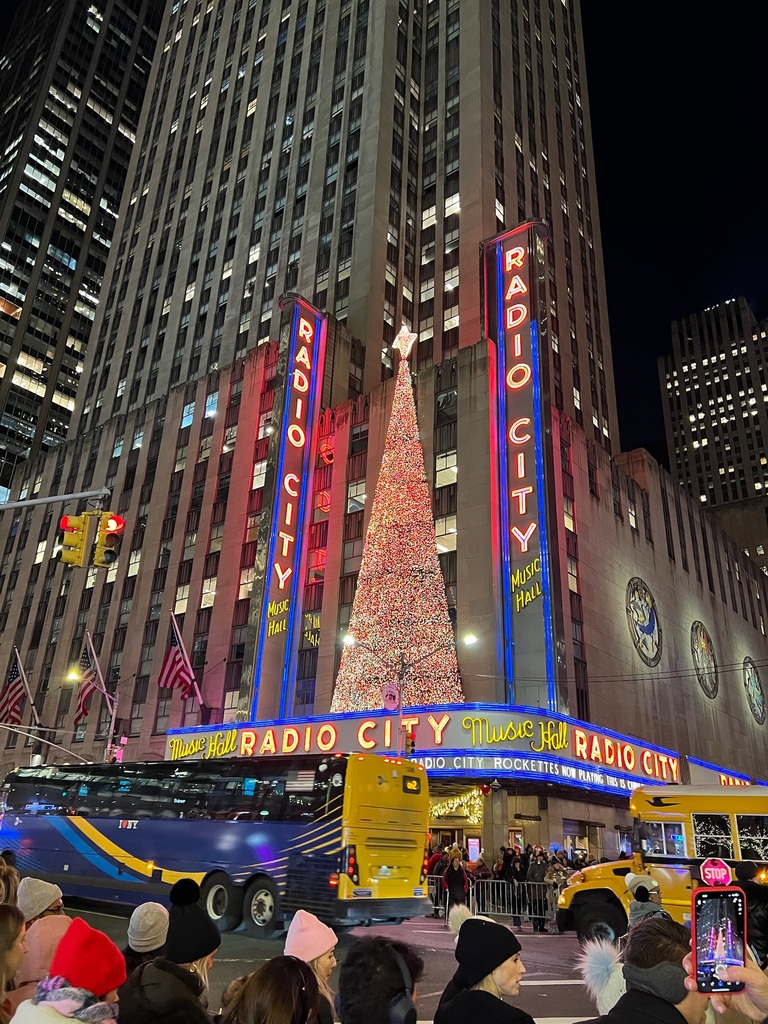 Street view of Radio City Music Hall at night, adorned with a sparkling Christmas tree. Neon red and blue signs glow, with bustling traffic and pedestrians.