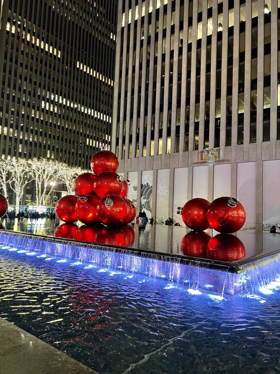 Large red Christmas ornaments are stacked on a reflective platform above a waterfall fountain, surrounded by illuminated trees and tall buildings at night.