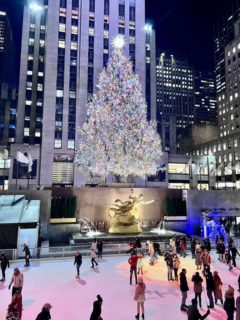 Illuminated Christmas tree with colorful lights at a city plaza, towering above a gold statue. People ice skate below, creating a festive atmosphere.
