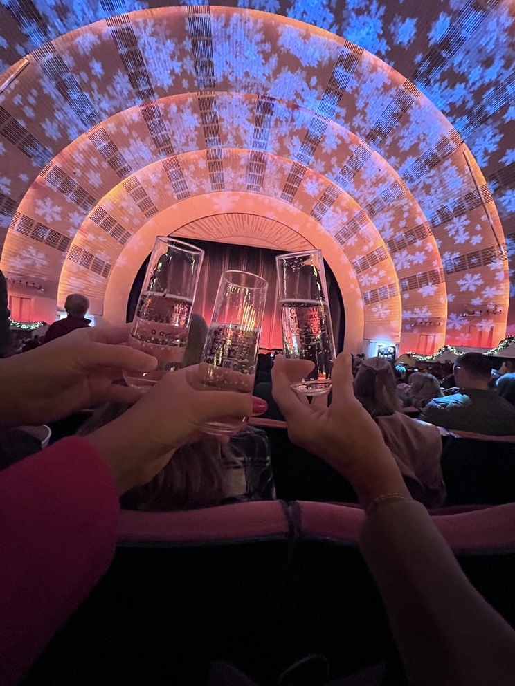 Hands hold up champagne glasses inside a large theater, with festive snowflake lights projected on orange arches, creating a celebratory atmosphere.