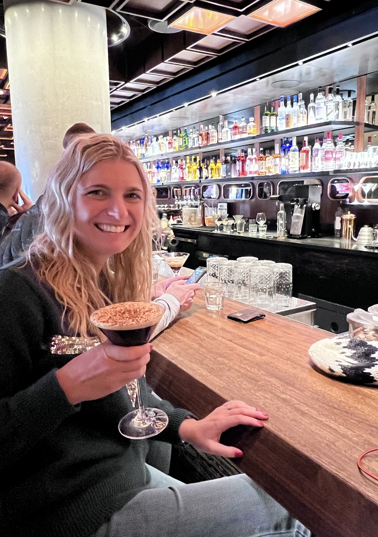 Woman with long blonde hair smiling and holding a cocktail at a bar. The bar features a variety of colorful bottles on a backlit shelf, creating a lively atmosphere.