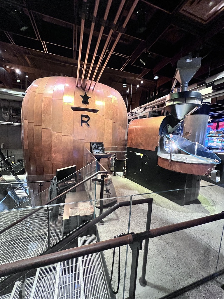 Copper coffee roasting equipment with industrial pipes and a large "R" symbol on a smooth, polished surface. The scene has an urban, modern vibe.