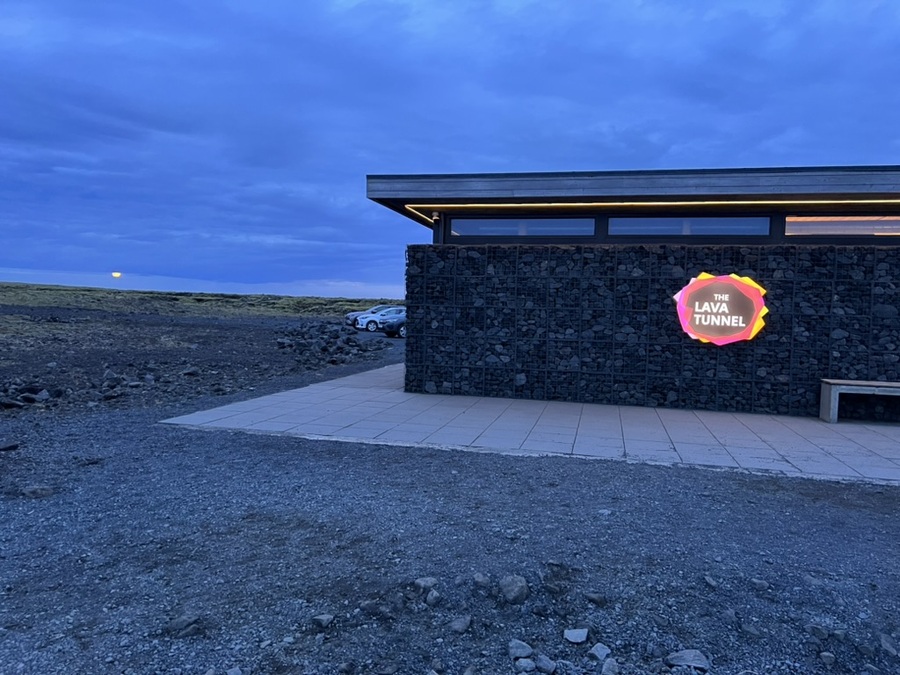 Building with a dark stone facade and a glowing sign reading "The Lava Tunnel" against a dusk sky. Sparse landscape and parked cars in the background.