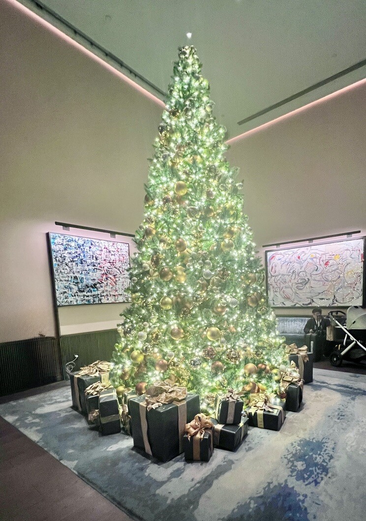 A tall, elegantly decorated Christmas tree adorned with golden ornaments and lights stands in a modern room. Wrapped gifts sit beneath it, creating a festive atmosphere.