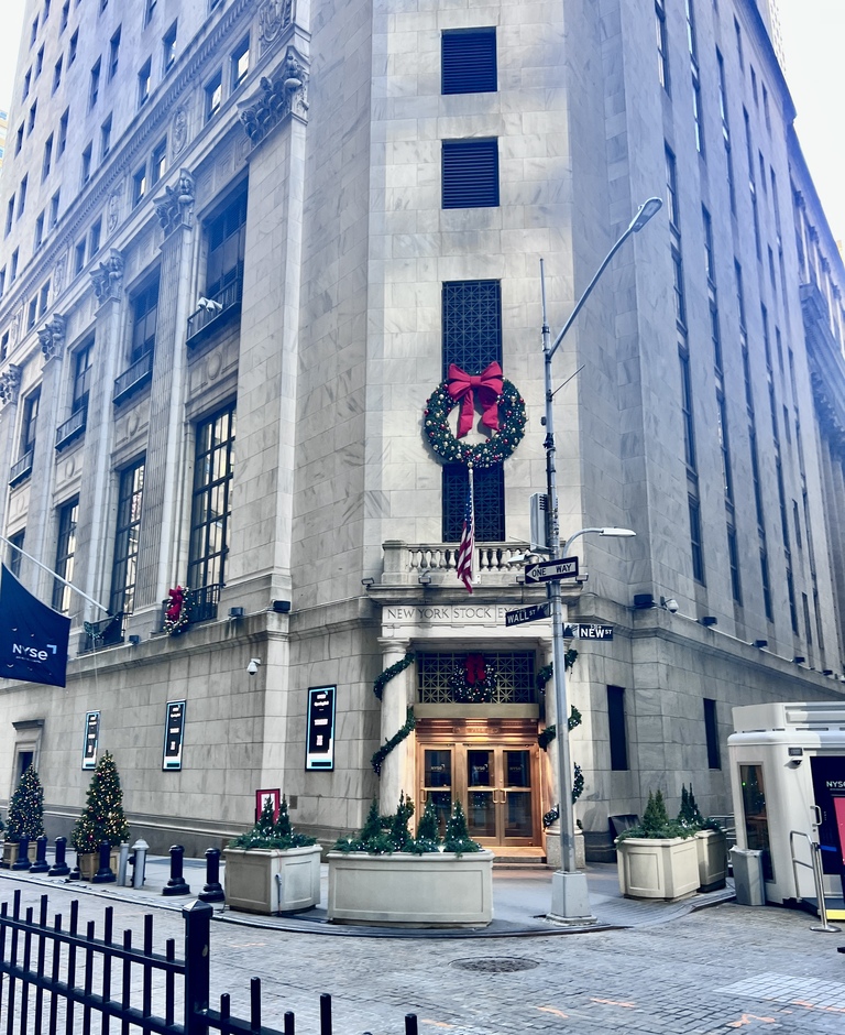 Street view of a tall building adorned with festive holiday decorations, including wreaths and garlands. A street sign reads "Wall St" and "New St."