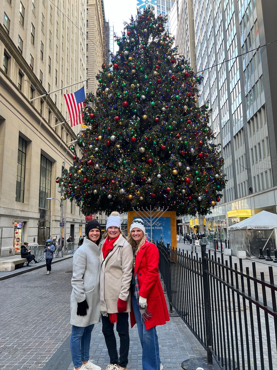 Three people in winter coats and hats smile in front of a large, decorated Christmas tree in an urban setting. American flag visible behind them. Festive atmosphere.