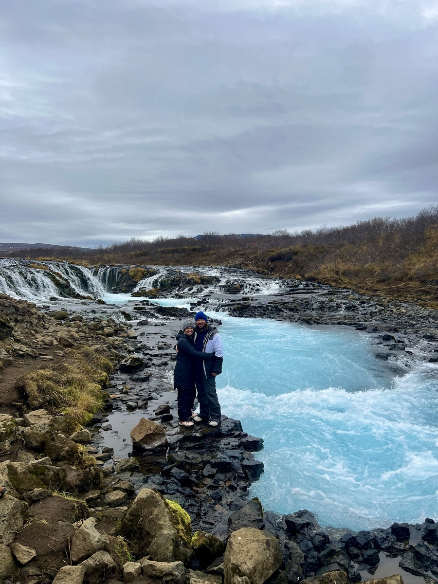 A couple embraces on rocky terrain by a vivid blue waterfall under an overcast sky. The scene conveys warmth amidst the chilly natural setting.