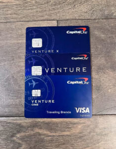 Three Capital One credit cards are stacked on a wooden surface. They are labeled Venture X, Venture, and Venture One. Each card has a dark blue design with travel-themed graphics.
