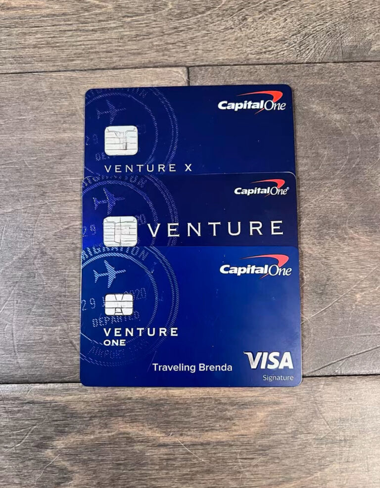 Three Capital One credit cards are stacked on a wooden surface. They are labeled Venture X, Venture, and Venture One. Each card has a dark blue design with travel-themed graphics.