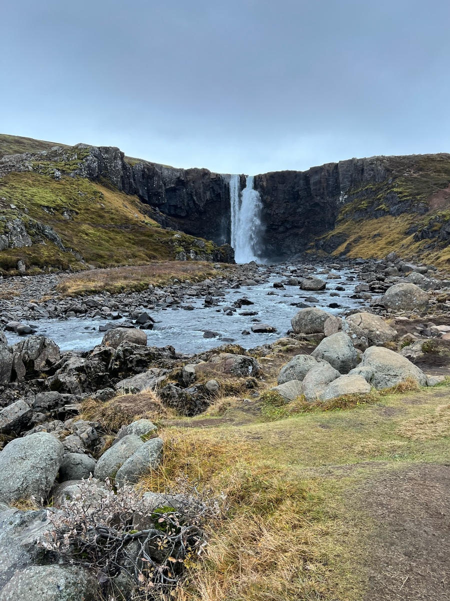 Majestic waterfall cascading over a rocky cliff into a serene river, surrounded by lush green hills and scattered boulders under an overcast sky.