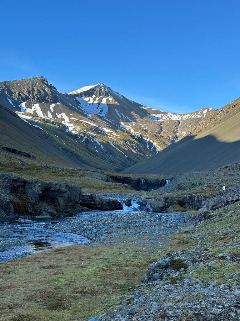 Snow-capped mountains under a clear blue sky, with a small waterfall flowing into a rocky stream. The scene is tranquil and picturesque.