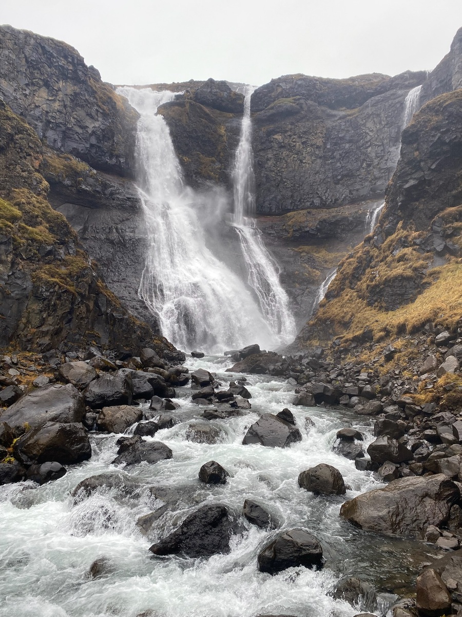 Rugged landscape with a multi-tiered waterfall cascading over dark rocks. Surrounded by autumnal grass, the water flows into a rocky stream below.