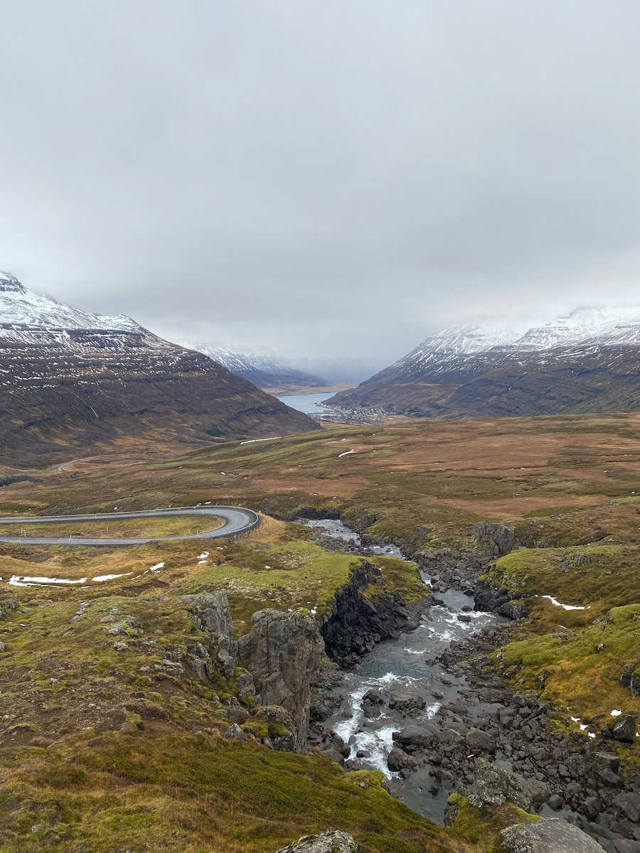 A winding river cuts through a rocky valley with moss-covered rocks, surrounded by snow-capped mountains under an overcast sky, evoking serenity.