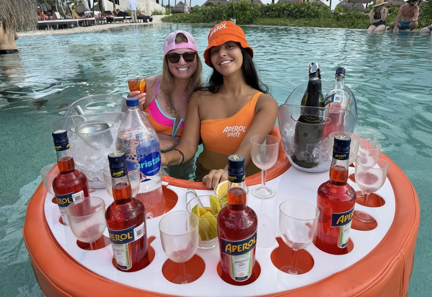 Two women in a pool smile behind a floating drink tray with Aperol bottles, lemon slices, and glasses. Vibrant poolside ambiance and leisure.