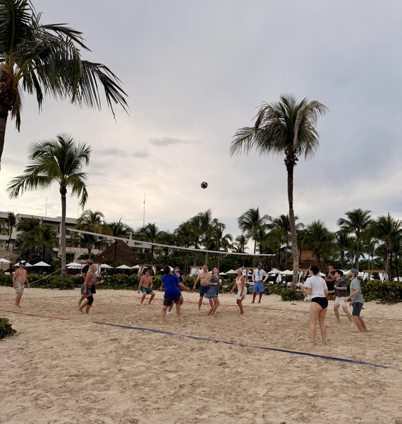 A group of people play beach volleyball under cloudy skies, surrounded by palm trees. The scene is lively and relaxed, evoking a sense of fun and leisure.