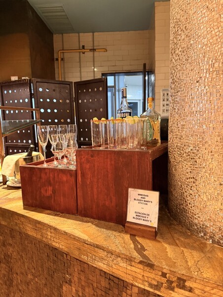 Buffet setup with wine glasses, citrus-garnished glasses, and alcohol bottles on a wooden stand. A sign reads "Mimosa & Bloody Mary Station." Warm ambiance.