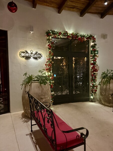 Elegant entrance to Bordeaux restaurant, adorned with festive garlands and lights. Large planters flank the dark metal doors, and a red-bench adds charm.