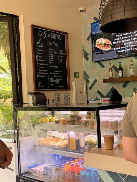 Coffee shop interior with menu board listing drinks like cappuccino and espresso. Display fridge shows sandwiches, muffins, and bottled drinks. Casual atmosphere.