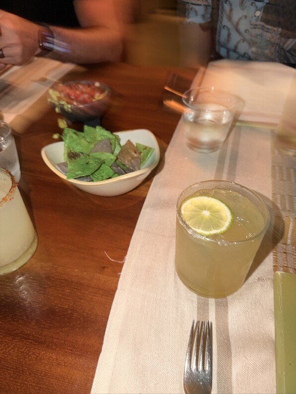 A table with a margarita, bowl of green and brown tortilla chips, salsa, and utensils. The setting is warm, suggesting a casual dining atmosphere.