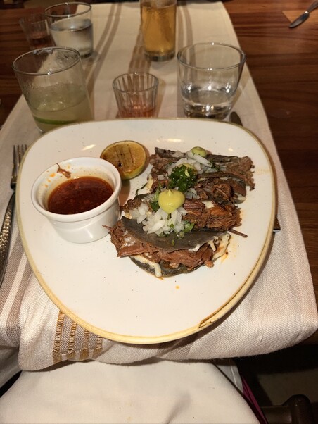 A plate of tacos with shredded meat, onions, and salsa, accompanied by a lime slice and a small dish of sauce. Drinks and glasses on a wooden table.