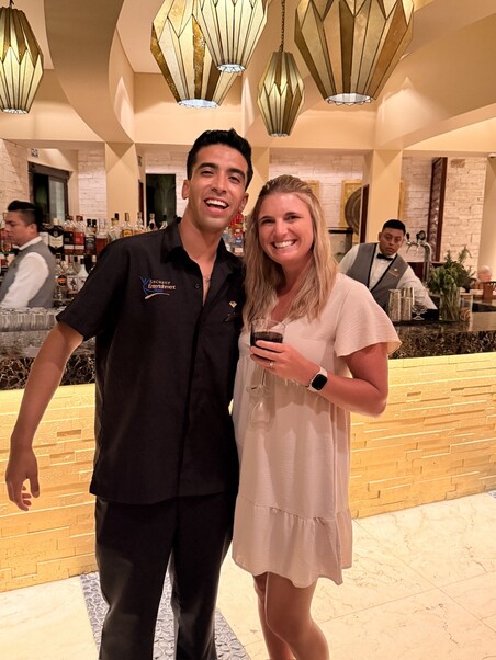 A smiling man and woman stand together in a warmly lit bar, holding drinks. Stylish pendant lights and bartenders are visible in the background.