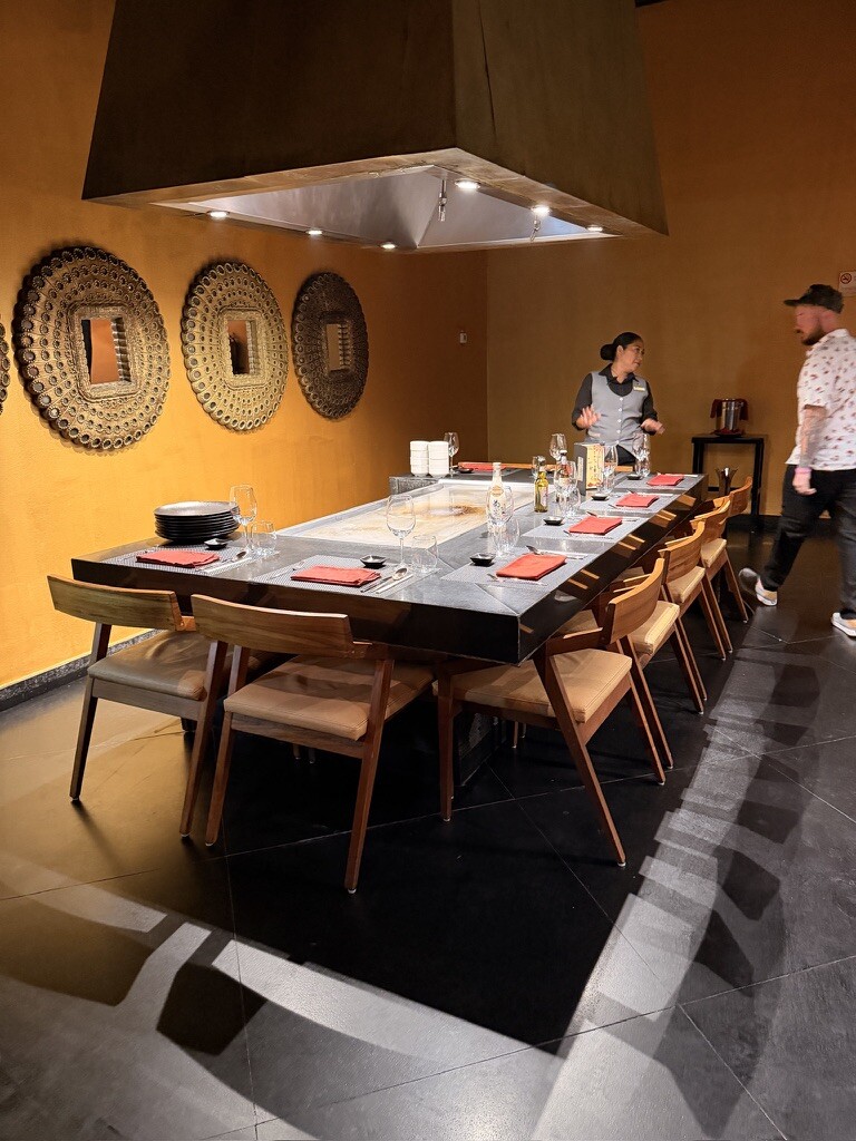 Modern dining room with a large rectangular table set for a meal, surrounded by wooden chairs with red napkins. Two people are present. Gold wall decor adds elegance.