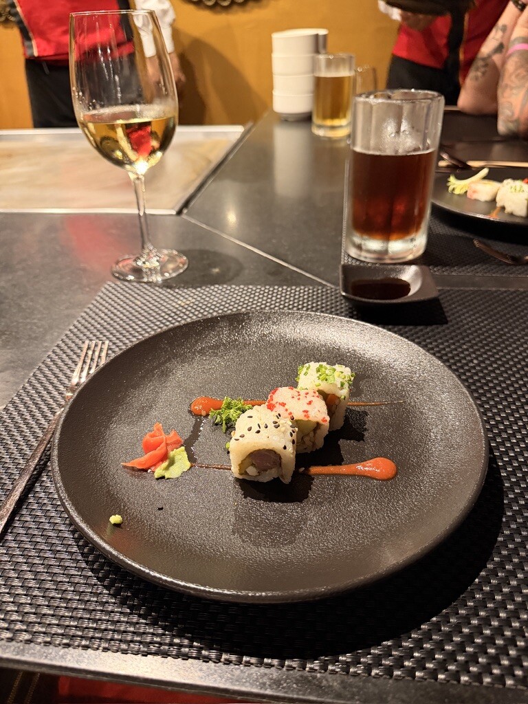Plate of sushi rolls with vibrant toppings on a textured black mat, accompanied by soy sauce, a glass of white wine, and iced tea. Elegant dining ambiance.