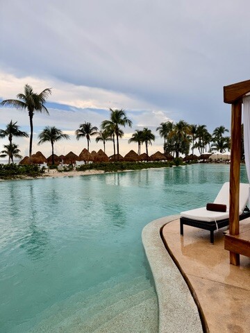 Relaxing tropical pool scene with palm trees, thatched umbrellas, and a lounge chair by the water. The serene sky suggests a peaceful atmosphere.