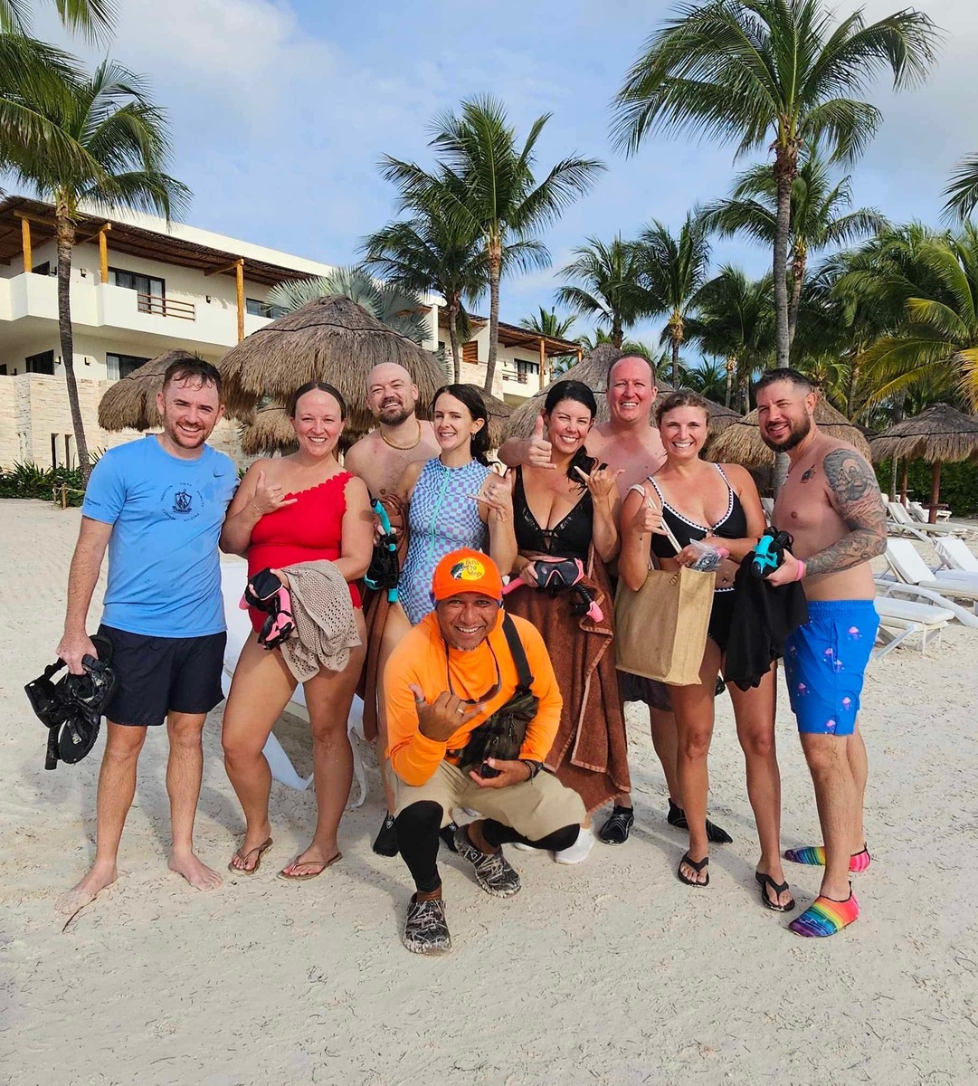 A group of nine people, smiling and posing on a sandy beach with palm trees and resort huts in the background. They are wearing swimsuits, holding snorkeling gear. The mood is joyful and relaxed.