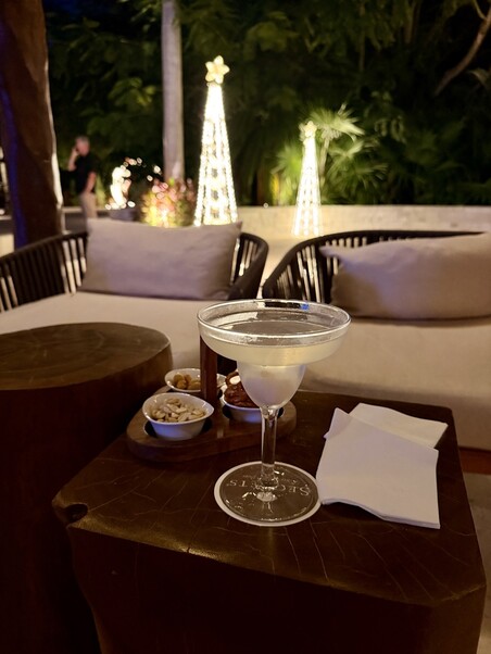 A martini glass on a wooden table with snacks, set in a tropical outdoor lounge at night. Illuminated Christmas trees in the background create a festive ambiance.