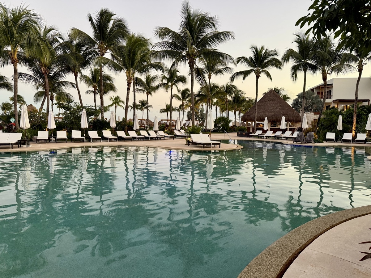 Tropical resort pool with calm water, surrounded by palm trees and lined with white loungers and umbrellas. A thatched hut and a serene atmosphere.