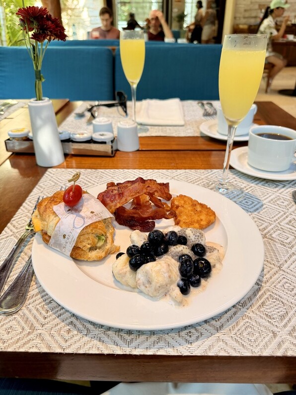 A breakfast plate on a wooden table features a croissant sandwich, crispy bacon, hash brown, and blueberries on cream. Two glasses of mimosa and a coffee cup are in the background, creating a cozy brunch scene.