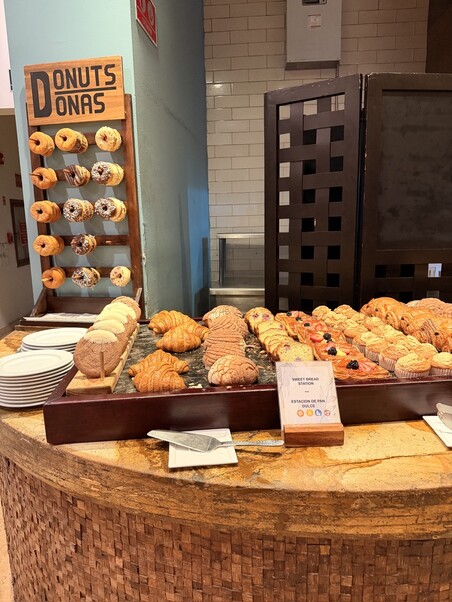 A wooden stand labeled "Donuts/Donas" displays assorted donuts. A counter holds croissants and pastries. Plates and napkins are ready on a rustic surface.