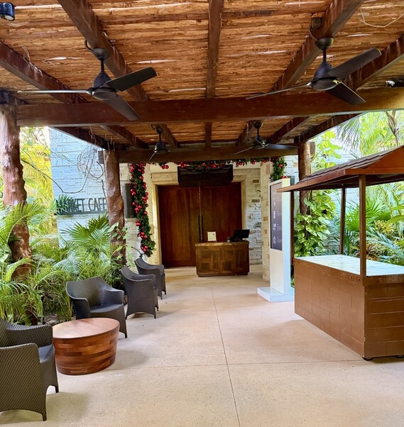 Covered outdoor cafe with wicker chairs and a wooden table on a tiled patio. Ceiling fans hang above, surrounded by lush greenery and festive garlands.