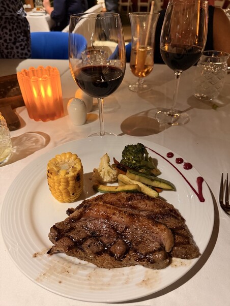 A gourmet dish featuring a juicy steak, grilled corn, and assorted vegetables on a white plate. Two glasses of red wine are in the background on a white tablecloth, with warm candlelight creating an elegant dining atmosphere.