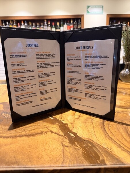 The image shows an open cocktail menu on a wooden bar table. The menu lists various cocktails and club specials. Shelves with bottles are blurred in the background.