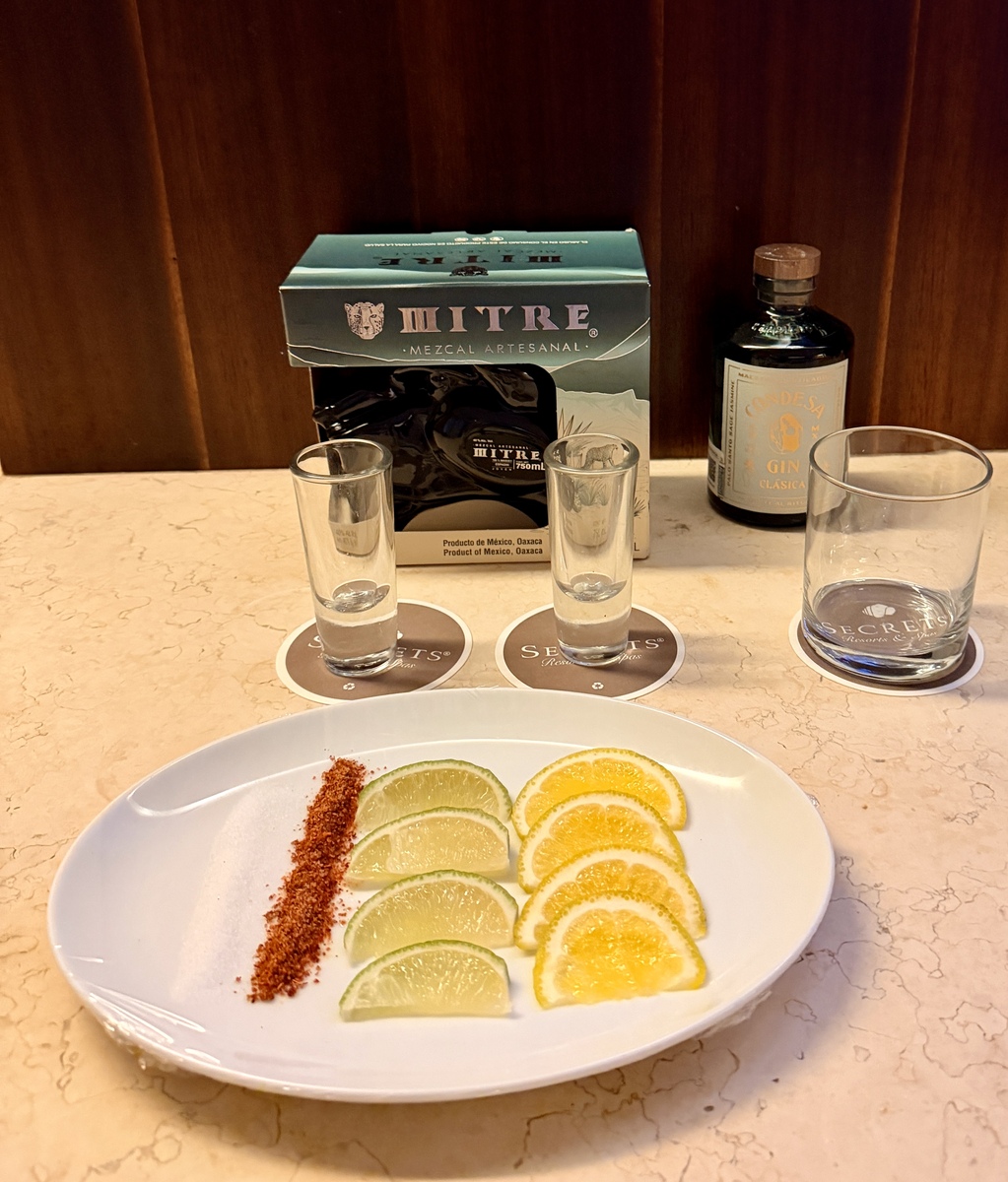 A serving setup with a Mitre Mezcal box, two shot glasses on coasters, a gin bottle, and a plate of sliced citrus with chili powder on a marble surface.