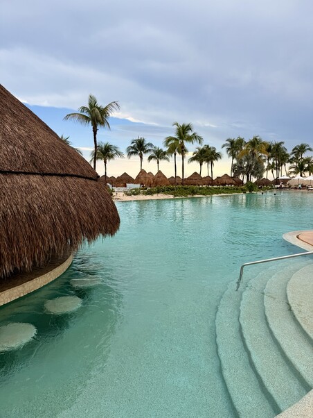 Serene poolside scene with turquoise water, stairs leading in, and a thatched-roof hut. Palm trees and loungers line the edge under a partly cloudy sky.