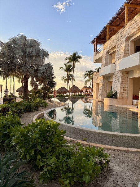 Luxurious resort at sunset, with a curved infinity pool, lush greenery, and tall palm trees. The stone architecture and tranquil setting evoke relaxation.