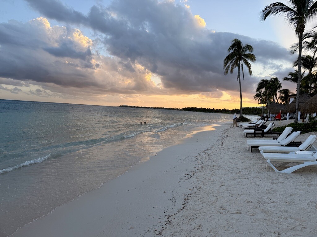Tropical beach at sunset with soft waves, white sand, and palm trees. Lounge chairs line the shore; warm, peaceful ambiance with vibrant clouds.