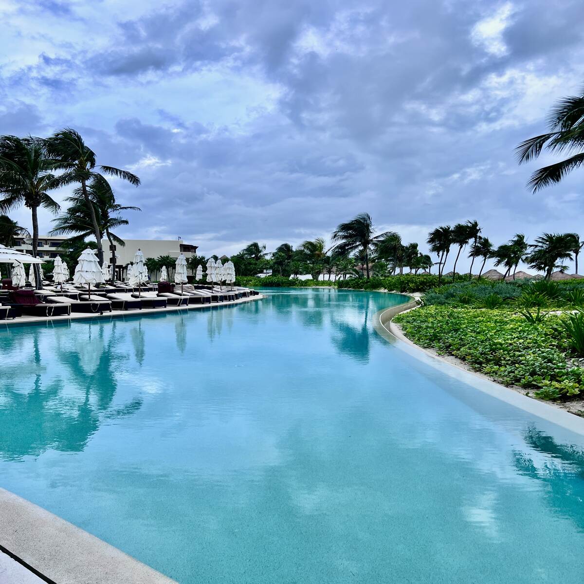 Infinity pool reflecting a cloudy sky, surrounded by lush greenery and palm trees. Lined with sun loungers and white umbrellas, conveying tranquility.