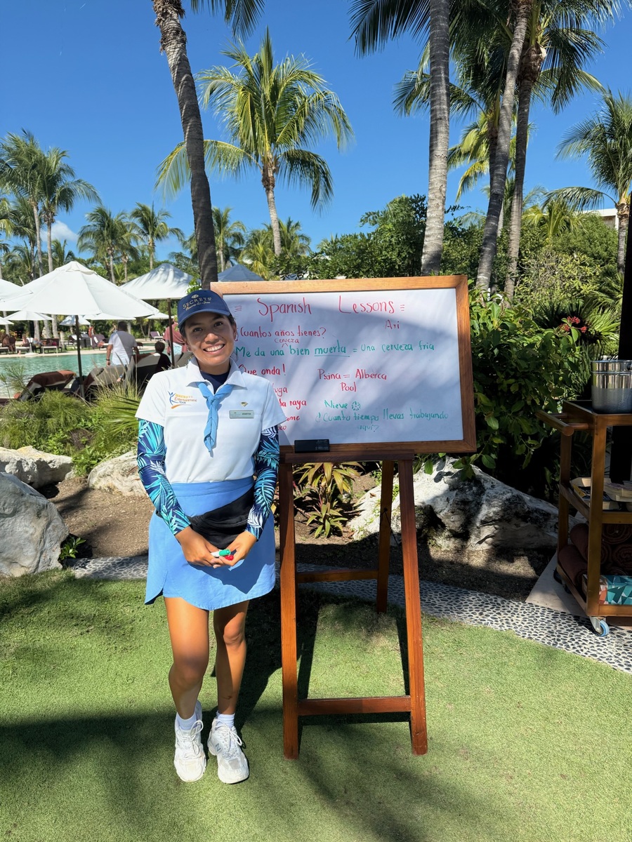 Woman smiling in a uniform stands beside an easel with a "Spanish Lessons" board, surrounded by palm trees and a pool. Bright, sunny day.
