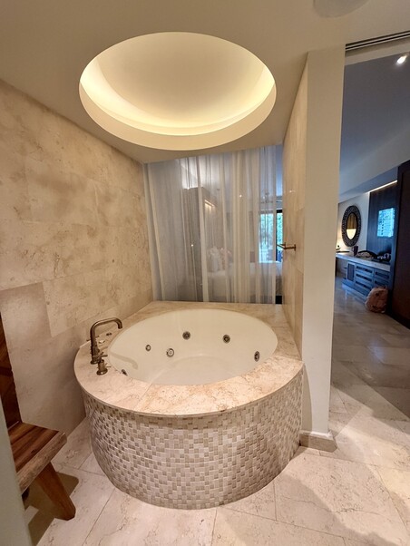 Luxurious bathroom with a round, tiled jacuzzi tub, soft lighting from a circular ceiling light, and sheer curtains, creating a serene ambiance.