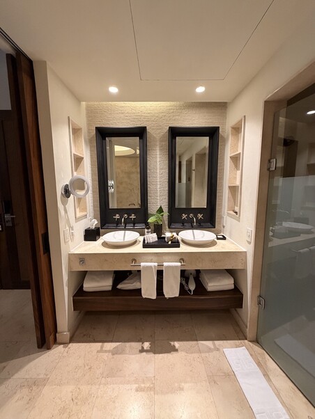 Stylish bathroom with two sinks, dark framed mirrors, and built-in shelves. Soft lighting and beige tiles create a calm, modern ambiance.